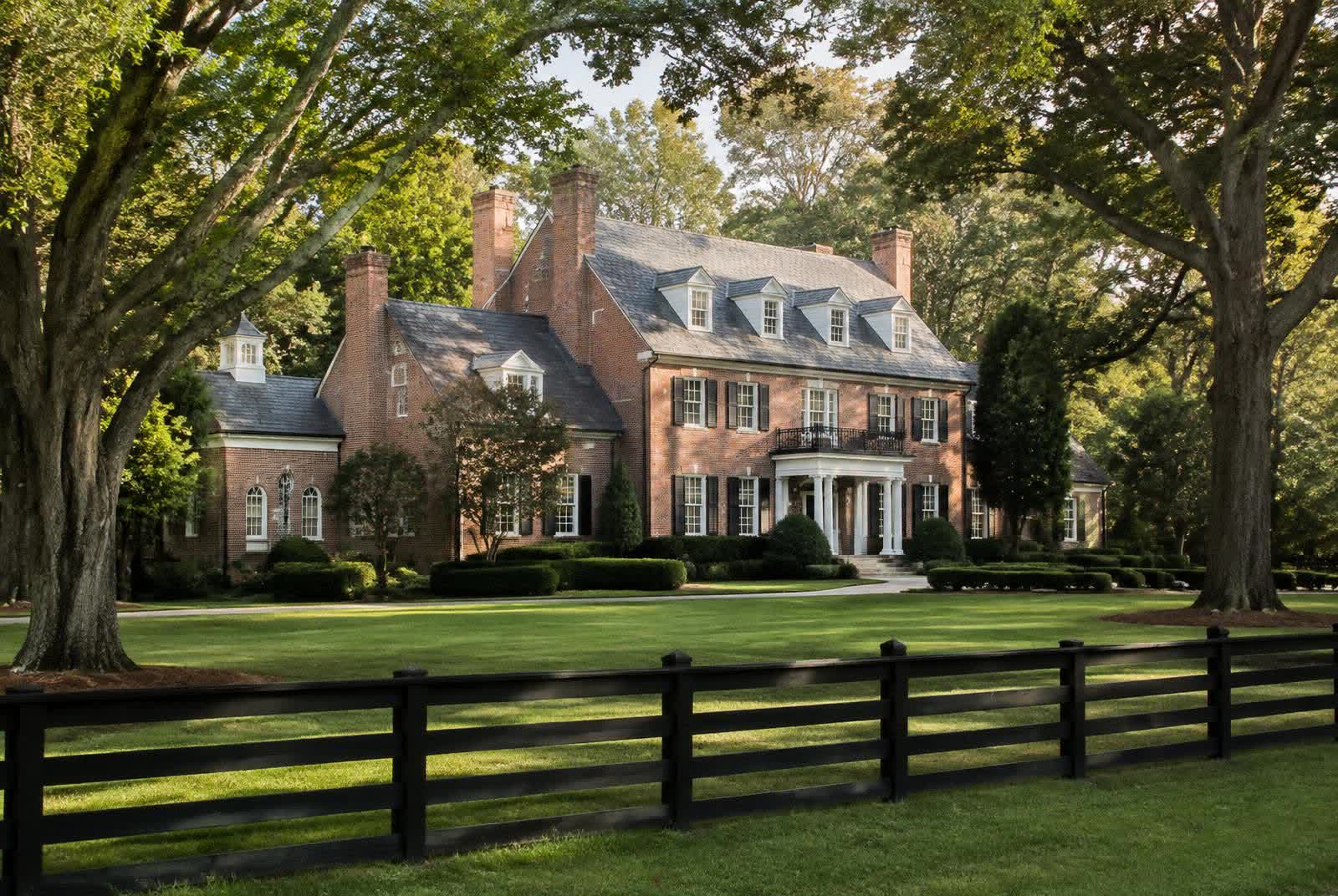 Traditional brick estate home with prominent chimneys behind black board fencing under mature oaks in Marvin, North Carolina